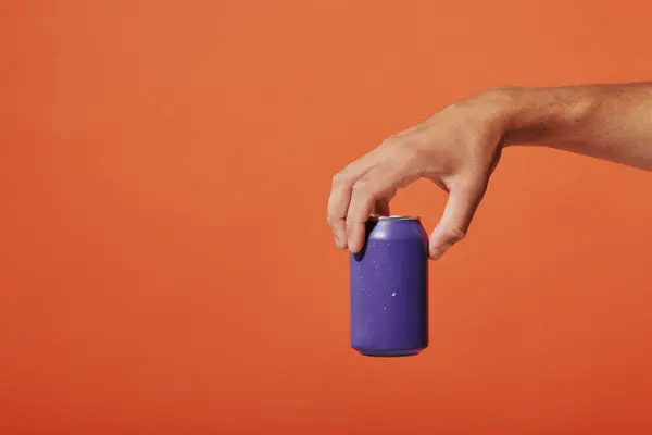 Vista recortada de la persona que sostiene lata de refresco púrpura en la mano sobre fondo naranja, bebida carbonatada - foto de stock
