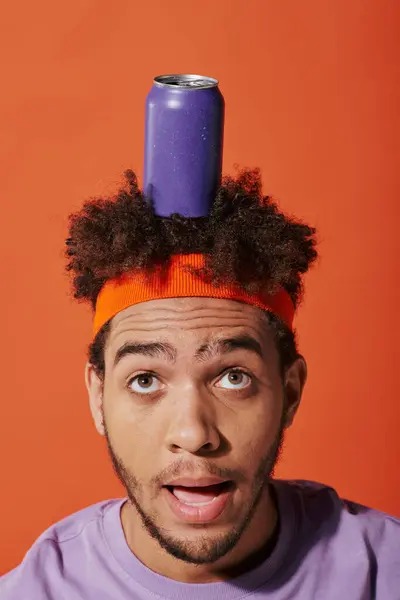 Purple soda can on head of shocked curly african american guy with headband on orange background — Stock Photo