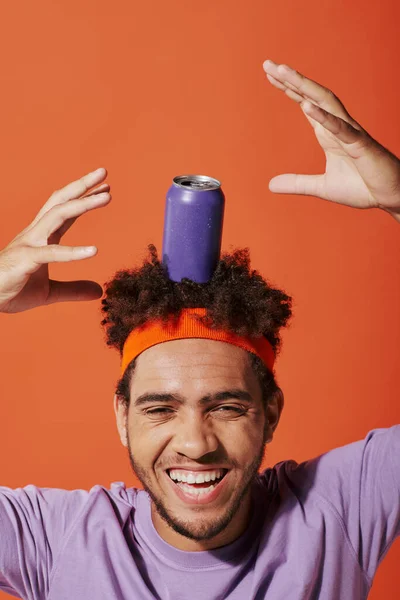 Purple soda can on head of happy curly african american man with headband on orange background — Stock Photo