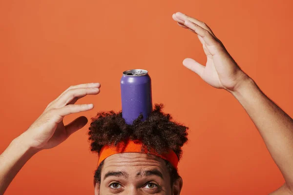 Purple soda can on head of curly african american man with headband on orange background, gesture — Stock Photo