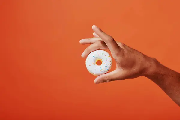 Tiro recortado de la persona que sostiene donut de vainilla vidriada con aspersiones sobre fondo naranja, glaseado blanco - foto de stock