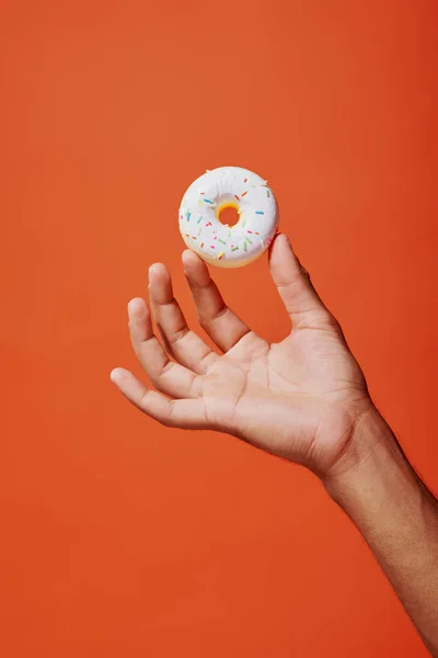 Tiro recortado de homem segurando donut de baunilha envidraçado com polvilhas sobre fundo laranja, glacê branco — Fotografia de Stock