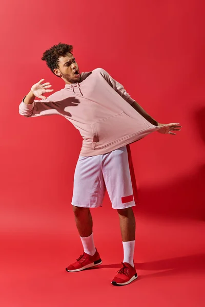 Chocado afro-americano homem olhando para roupas de ginástica e de pé com boca aberta no fundo vermelho — Fotografia de Stock