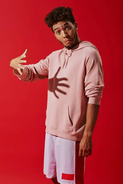 Confused african american man in hoodie gesturing and looking at camera on red background, shadow — Stock Photo