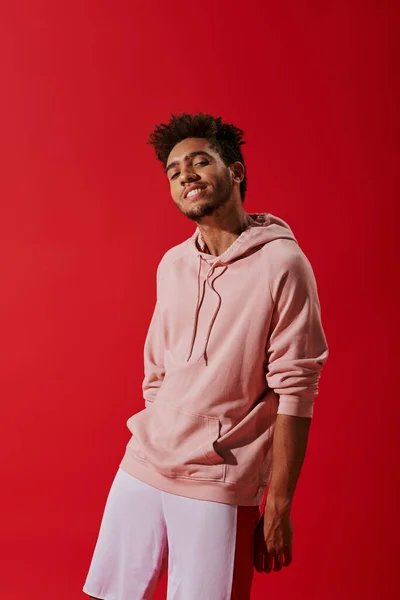 Happy african american man in pink hoodie looking at camera while smiling on red background — Stock Photo