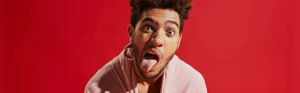 African american man pulling pink hoodie and sticking out tongue while on red background, banner — Stock Photo