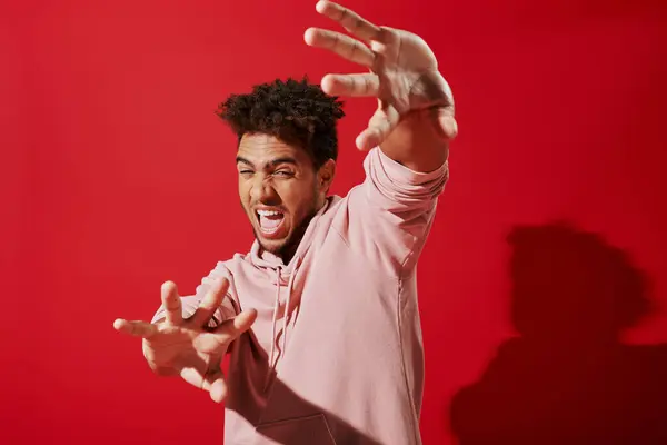 Ansioso afro-americano homem com capuz rosa gesticulando e gritando em fundo vermelho, aterrorizado — Fotografia de Stock