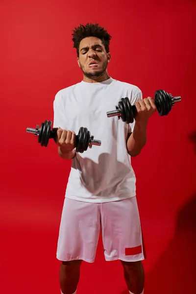 Fuerte afroamericano deportista haciendo ejercicio con pesadas mancuernas sobre fondo rojo, mueca - foto de stock