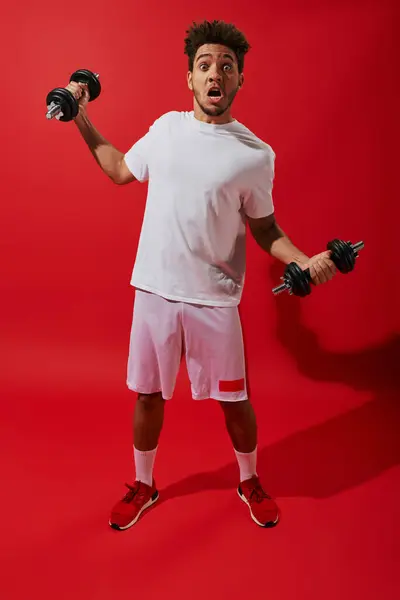 Shocked african american sportsman working out with heavy dumbbells on red background, grimace — Stock Photo