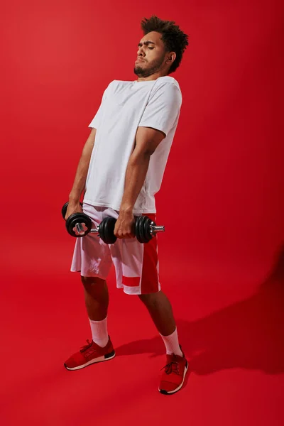 Sportif afro-américain bouclé travaillant avec de lourds haltères sur fond rouge, puissance — Photo de stock
