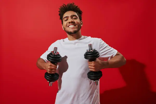 Alegre afroamericano deportista haciendo ejercicio con pesadas mancuernas sobre fondo rojo, poder - foto de stock