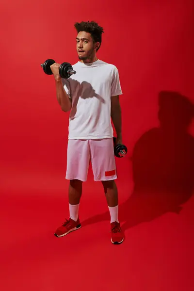 Full length of african american sportsman working out with heavy dumbbells on red background, power — Stock Photo
