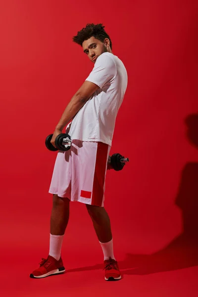 Full length of african american sportsman exercising with heavy dumbbells on red background, power — Stock Photo