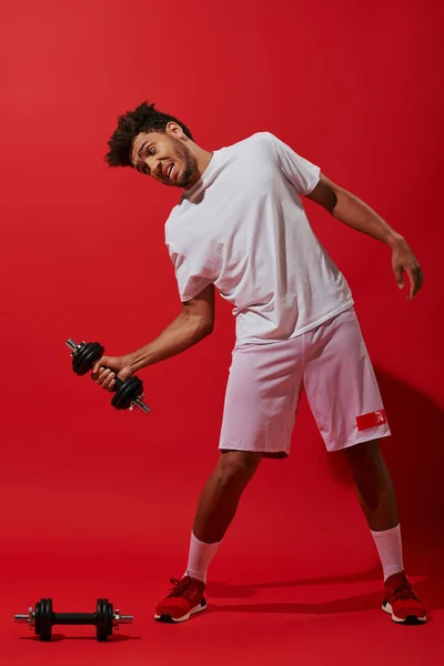 Longitud completa del hombre americano africano divertido en ropa deportiva que trabaja hacia fuera con la mancuerna sobre fondo rojo - foto de stock