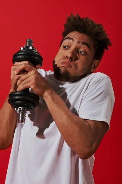 Young african american man working out with heavy dumbbell and puffing cheeks on red background — Stock Photo