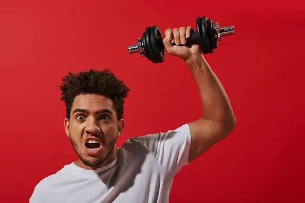 Homme afro-américain motivé en vêtements de sport travailler avec haltère lourde sur fond rouge — Photo de stock