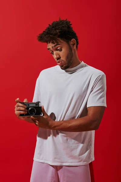 Joven afroamericano hombre mirando su cámara vintage sobre fondo rojo, fotografía como hobby - foto de stock