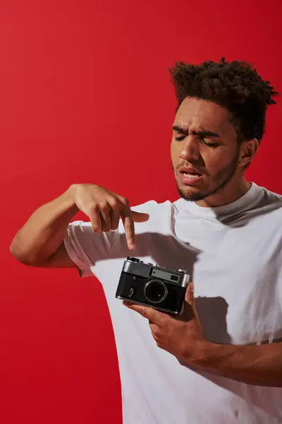 Jeune homme afro-américain regardant sa caméra vintage sur fond rouge, en appuyant sur le bouton — Photo de stock