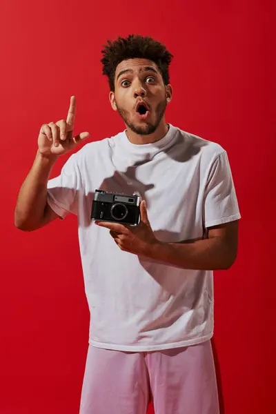 Joven afroamericano hombre teniendo idea mientras sostiene su cámara vintage sobre fondo rojo - foto de stock