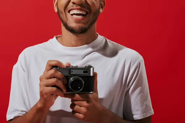 Fotografo afro-americano cortado e feliz segurando câmera vintage e sorrindo no fundo vermelho — Fotografia de Stock