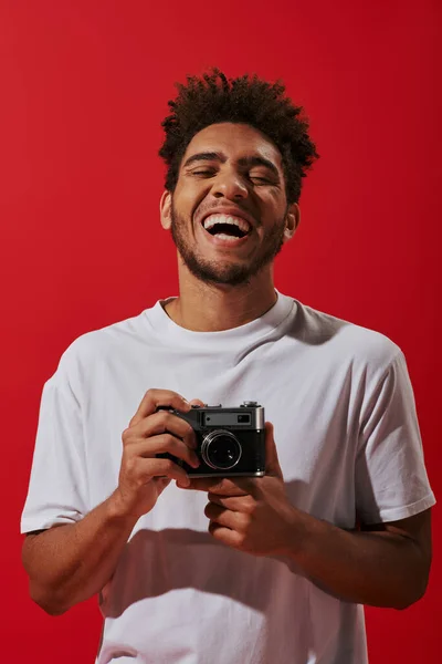 Alegre fotógrafo afro-americano segurando câmera vintage e sorrindo no fundo vermelho — Fotografia de Stock