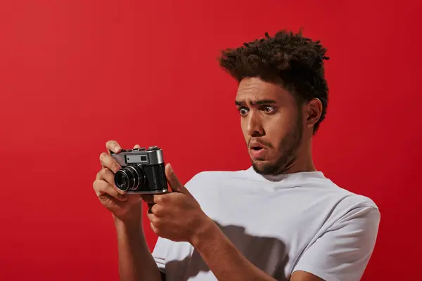 Funny african american photographer looking at retro camera while taking shot on camera on red — Stock Photo