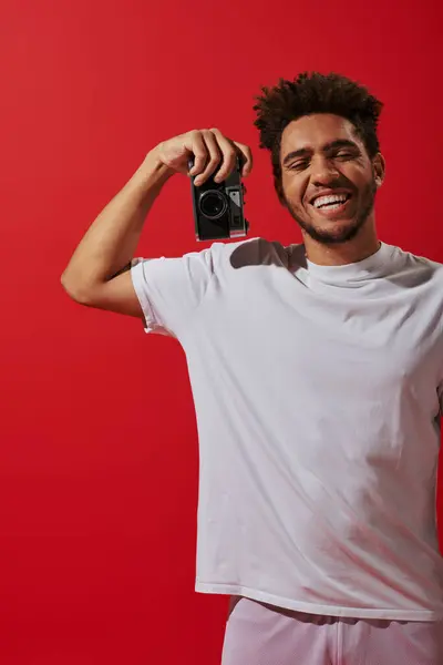Cheerful african american photographer holding retro camera and smiling on red background — Stock Photo