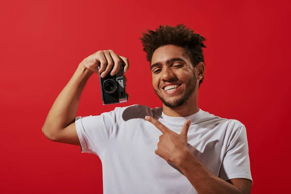 Веселый африканский американский фотограф указывает на ретро-камеру и улыбается на красном фоне — стоковое фото