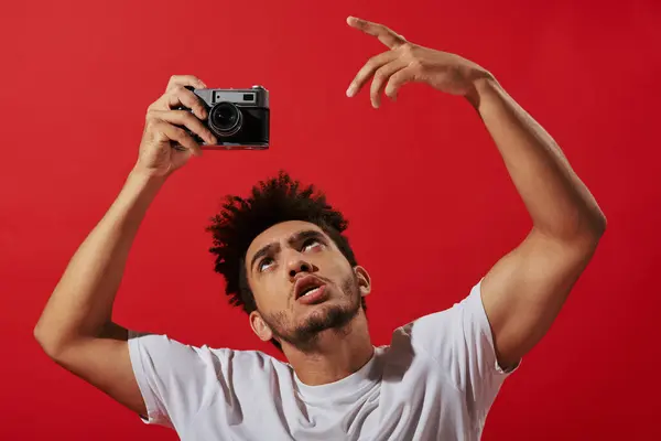 Смешной и творческий африканский американский фотограф, снимающий ретро-камеру на красном фоне — стоковое фото