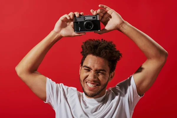 Joyful african american photographer holding retro camera and smiling on red background — Stock Photo
