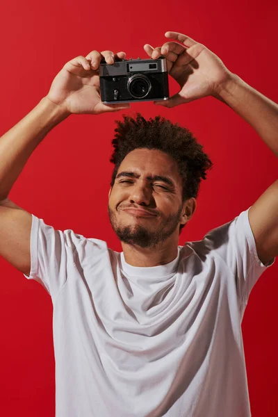 Funny african american photographer taking shot on retro camera and smiling on red background — Stock Photo