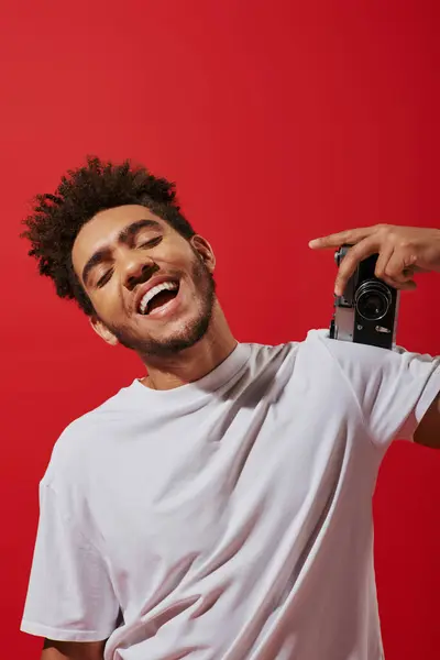 Optimistic african american photographer taking shot on retro camera and smiling on red background — Stock Photo