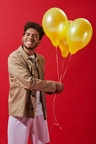 Cheerful african american man in beige jacket holding helium balloons on red background, party — Stock Photo