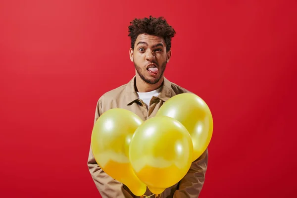 Funny african american man in beige jacket holding balloons and sticking tongue on red background — Stock Photo