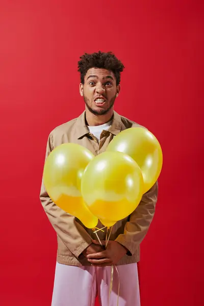 Funny african american man in beige jacket holding balloons and grimacing on red background — Stock Photo