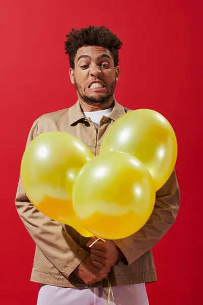 Funny african american man in beige jacket looking at balloons and grimacing on red background — Stock Photo
