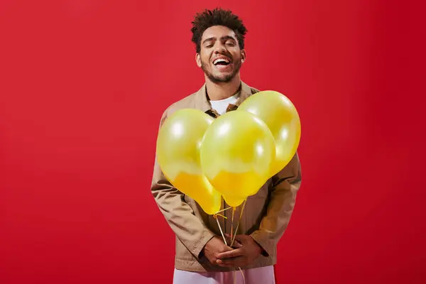 Cheerful african american man in beige jacket holding balloons and laughing on red background — Stock Photo