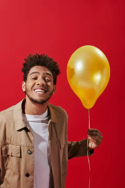 Positive african american man in beige jacket holding balloon and smiling on red background — Stock Photo
