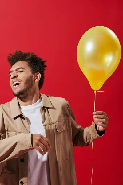 Optimistic african american man in beige jacket holding balloon and smiling on red background — Stock Photo