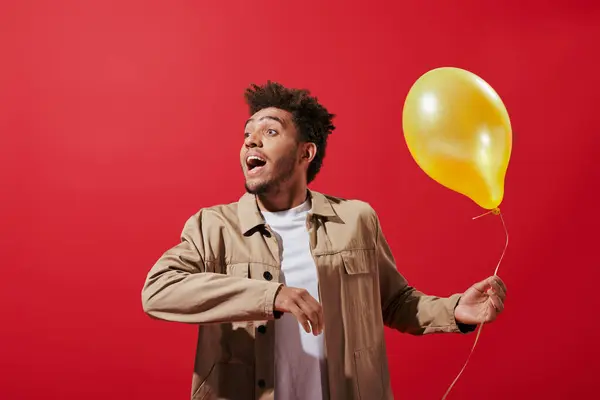 Joyful african american man in beige jacket holding balloon and smiling on red background — Stock Photo