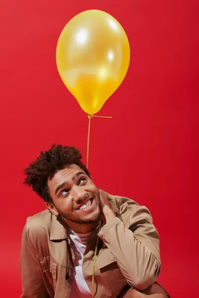 Pleased african american man in beige jacket smiling near yellow balloon on red background — Stock Photo