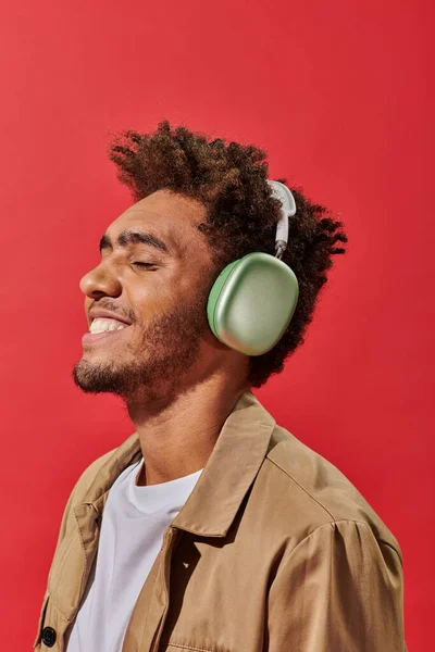 Portrait of happy african american man in wireless headphones listening music on red background — Stock Photo