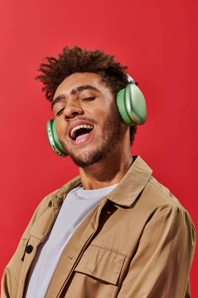 Portrait of excited african american man in wireless headphones listening music on red background — Stock Photo