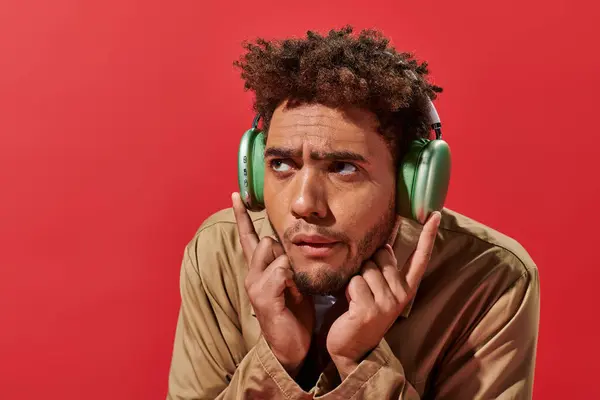 Portrait of confused african american man in wireless headphones listening music on red background — Stock Photo