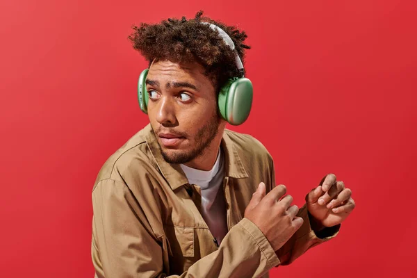 Portrait de l'homme afro-américain confus dans des écouteurs sans fil regardant loin sur fond rouge — Photo de stock