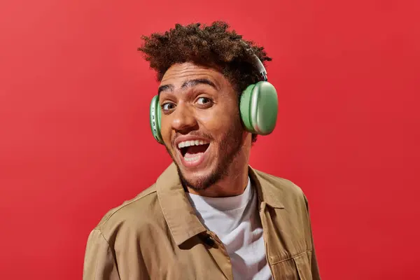 Porträt eines aufgeregten afrikanisch-amerikanischen Mannes in drahtlosen Kopfhörern mit offenem Mund auf rotem Hintergrund — Stockfoto