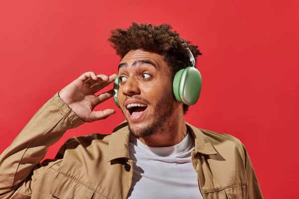 Portrait of amazed african american man in wireless headphones with open mouth on red background — Stock Photo