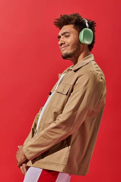 Portrait of pleased african american man in wireless headphones enjoying music on red background — Stock Photo