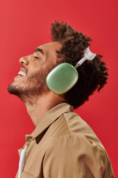 Vista lateral do homem americano africano alegre em fones de ouvido sem fio desfrutando de música no fundo vermelho — Fotografia de Stock
