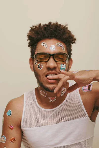 Young african american man in sunglasses with trendy stickers on face biting finger on grey backdrop — Stock Photo